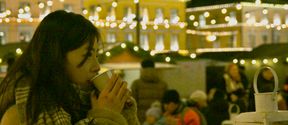 A woman wearing a big scarf is holidng a cup on her lips. Lights, buildings and people on the background.