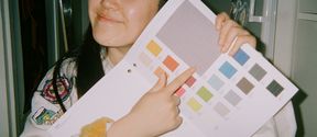 Person holding a colour swatch booklet and pointing at one of the swatches. They are wearing student overalls with colourful patches.
