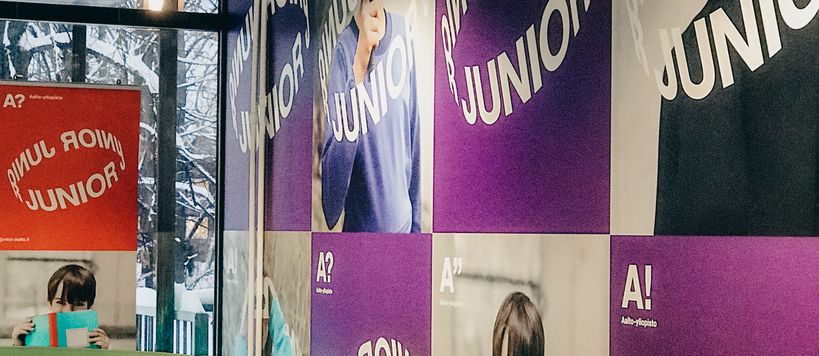 Aalto-yliopisto Juniorin aulatila: värikäs julisteiden peittämä seinä ja vihreä kulmasohva