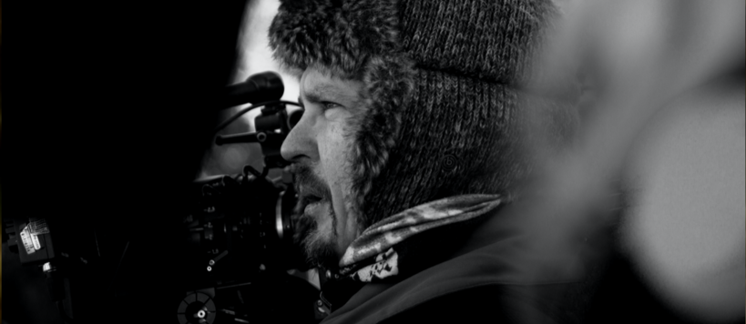 Mies kuvaa elokuvakameralla talvella ulkona, lämmin talvihattu päässään ihmisten seassa. Hänestä näkyy lähikuvassa vain profiili.