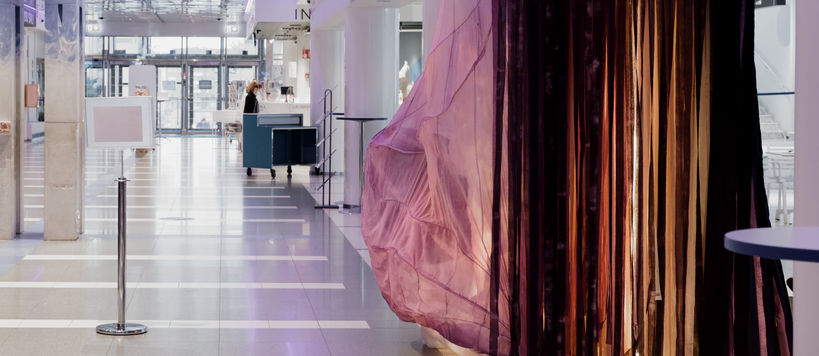 Kuvassa taideinstallaatio SOONI vaaleassa aulatilassa. Teoksen värikkäät tekstiilit liikkuvat teoksesta kantautuvan äänivirran mukana.