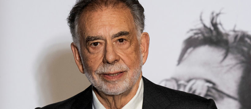 Harmaahiuksinen vanhempi mies puvuntakki ja kravatti yllään katsoo tummilla silmillään suoraan kameraan, taustalla näkyy valokuvaseinä.