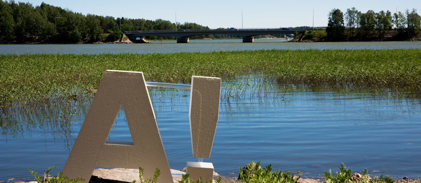 A white, 80 cms high 3D logo with capital A and an exclamation mark to the right of it. The logo is standing in the sunshine on a beach with the sea and some seagrass behind it. Picture by Aalto University / Mikko Raskinen 