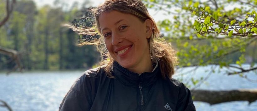 Carolina Weigl alumna from Aalto University Summer School course Nordic Biomaterials with CHEMARTS sitting in the nature by a lake in an outdoor jacket smiling directly into the camera.