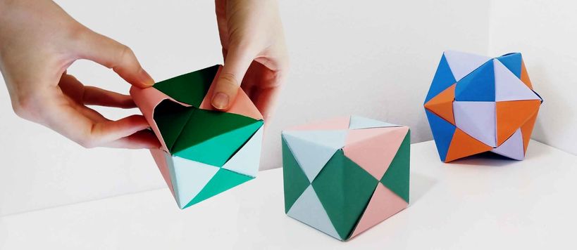 Kädet taittelevat värikästä origamikuuutiota, taustalla origamikuutio sekä tähtimäistetty oktaedri