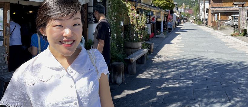 Chisako Ogiwara Digital Business Master Class alumna from Aalto University Summer School standing outside a restaurant at a street with some lush and green mountains in the background. She is wearing a short-sleeved white blouse and is smiling towards the camera.