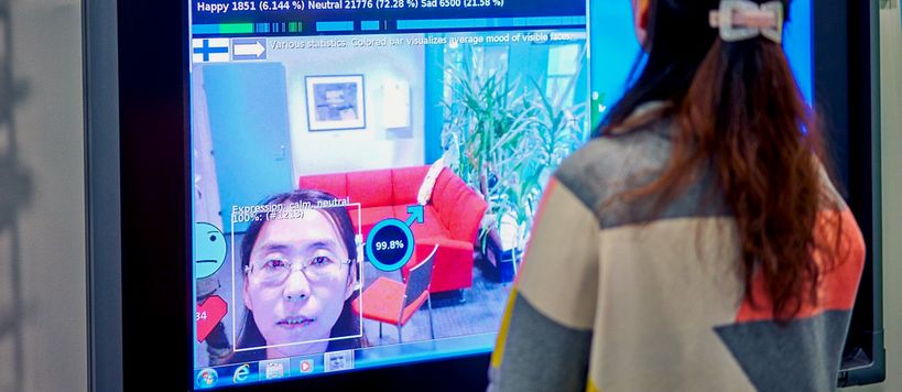 Professor Guoying Zhao, from the University of Oulu, standing in fron of a smart TV, showing a face analysis demo.
