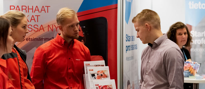 Three people with red shirts are standing in front of a roll-up with Hilti logo and a man in grey shirt is standing in front of them and looking at a broschure stand