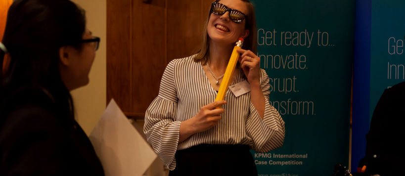 A woman is standing in front of roll-ups with KPMG logo, wearing sunglasses, smiling and holding a large pen next to her face.