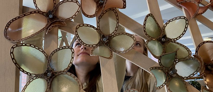 Two young people are looking upwards and smiling with mouths closed, behind an installation they designed. Art is made of wood and biopolymer flowers.