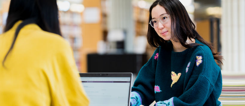 Two students studying by a laptop in Aalto University School of Arts, Design and Architecture. The student in focus is wearing a green knitted sweater and is looking insightfully at the other, who's back is positioned towards the camera.