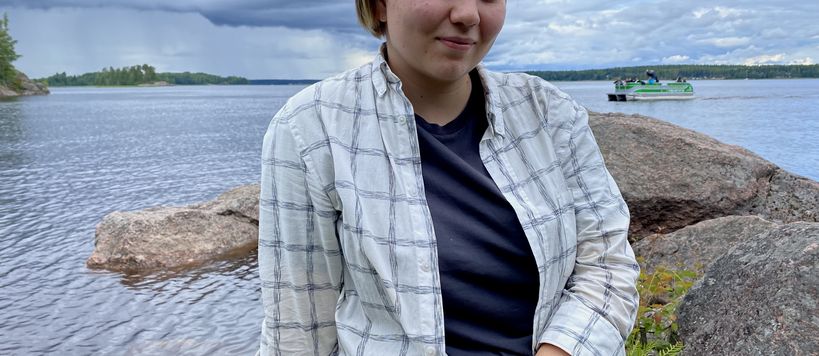 Elizaveta sitting on a rock at the seaside. There is a small green ferry in the background.