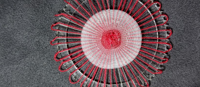 Kuvassa pyöreä kangas, joka on kudottu säteittäin ympyrän muotoon. Väreinä valkoinen ja punainen.