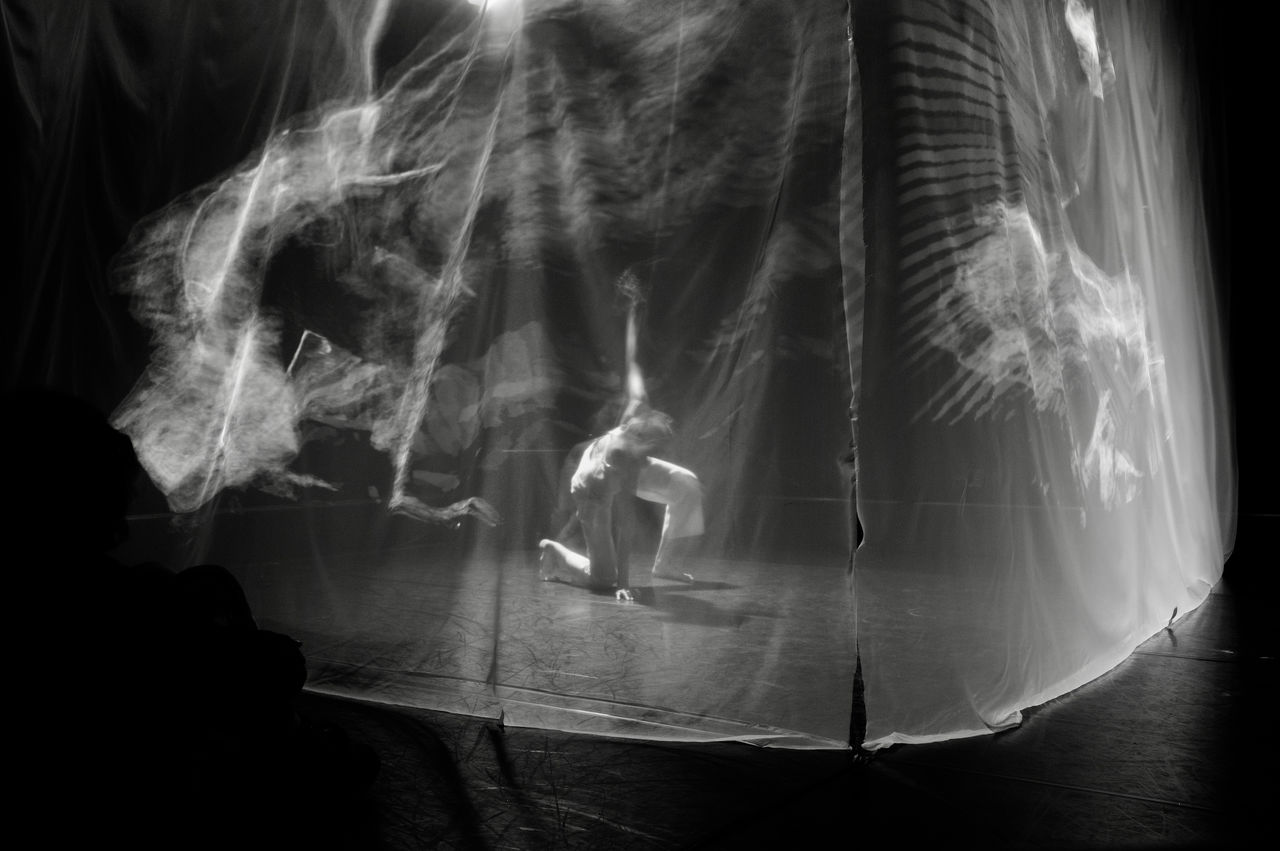 black and white photograph of artist Yo-Yo Lin performing on a stage with right knee and her left hand touching the floor. She is surrounded by a translucent scrim with graphical images projected upon it. 