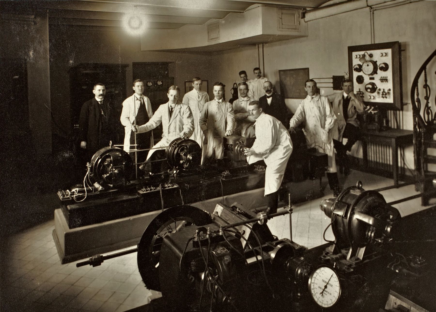 Historiallisessa kuvassa ryhmä miesopiskelijoita ja heidän opettajansa poseeraavat suurten mekaanisten koneiden äärellä