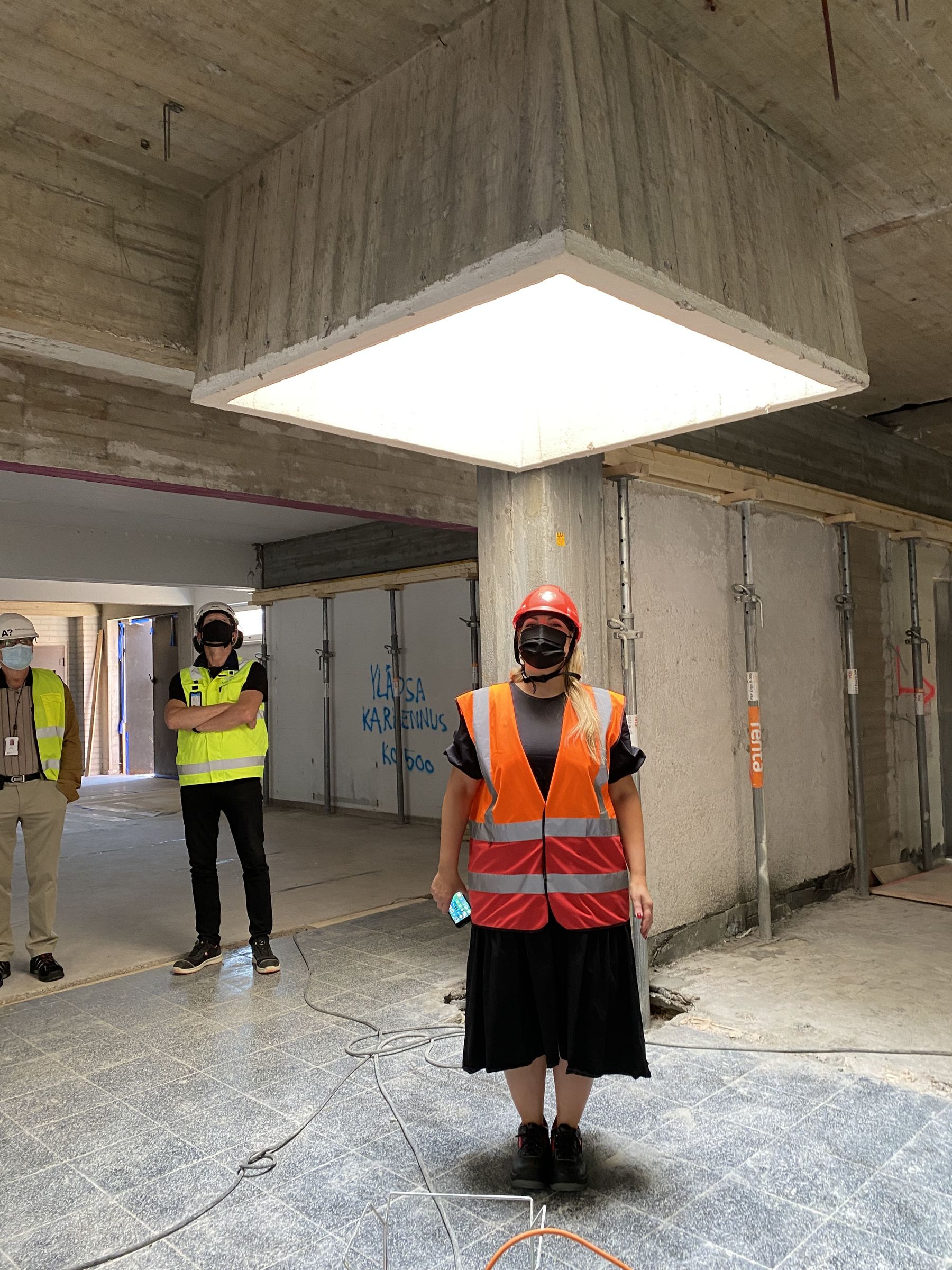 Two people look towards the third person standing in the middle of the picture, facing the camera. She wears a black dress and is under a skylight inside a building under construction