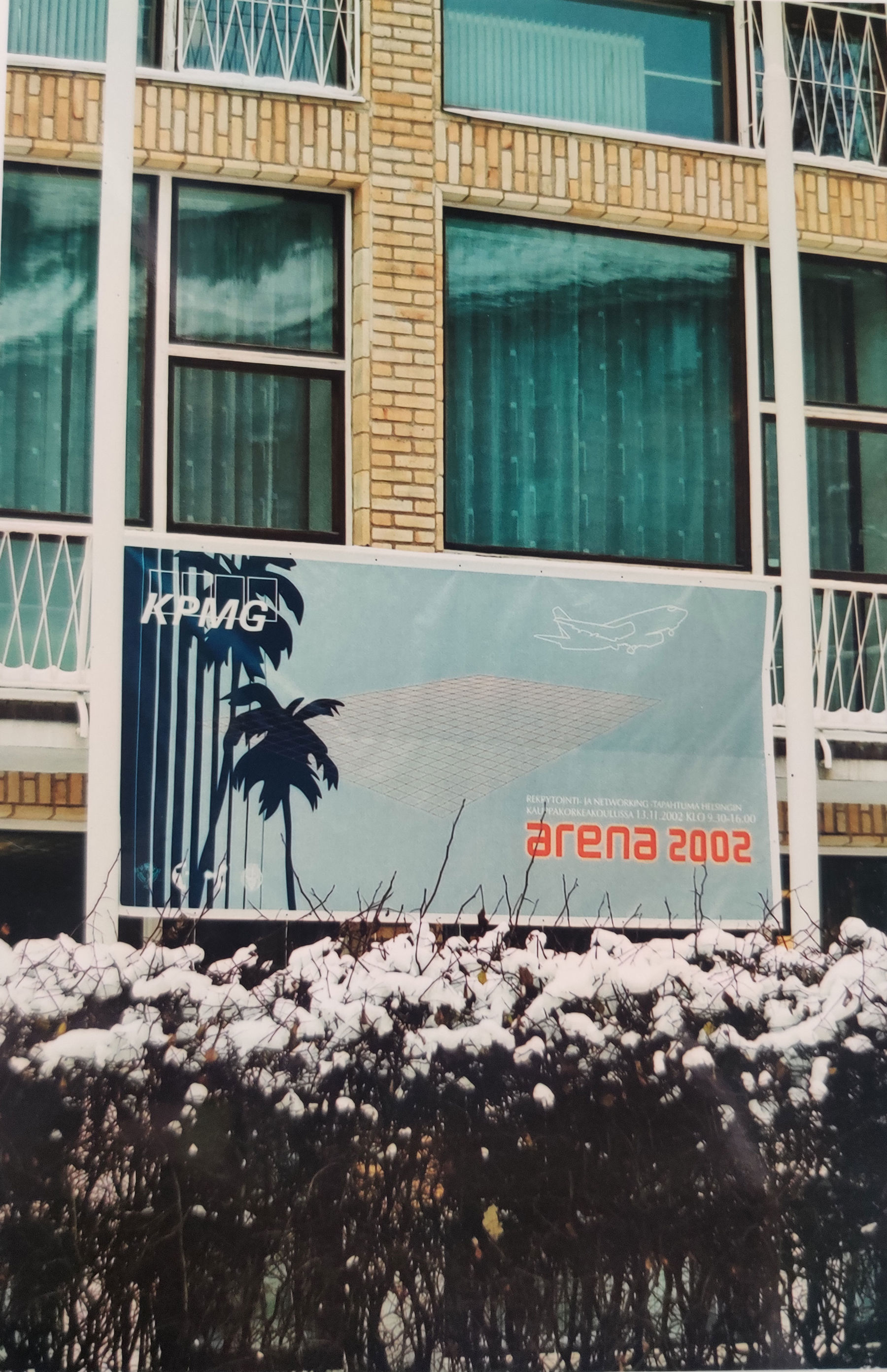 Picture of a building wall, with a billboard ad of an airplane, palm trees and texts KPMG and ARENA 2002.