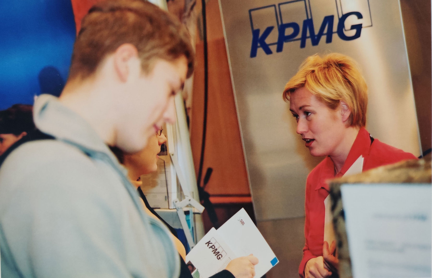 An old photo of two people close up. One is reading a broschure and another has her mouth open. KPMG logo in the background.