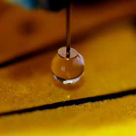 On image center, a spherical water droplet hangs from the tip of a disk, at the end of a sensor probe. Underneath lies a colourful yellow butterfly wing with black stripes, slightly out of focus toware the top and bottom of the image. Small individual scales that compose wing are visible.