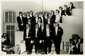 Mustavalkoisessa kuvassa juhlapukuisia miehiä seisoo portaikossa.