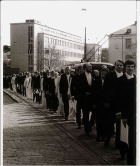Mustavalkoisessa kuvassa juhlavaatteisiin pukeutuneet promootiovieraat kulkevat kadulla kulkueessa.