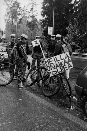  Capture 1992. Cavemen in a capture process stationed next to the bicycles of the Finnish Army (SA-INT). The text on the sign reads: "We have time to tread." Crafted miniature flag with bars with the Cave logo. Photo: TKY Archive, AYY Archive 