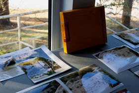 Photographs of the coastline lay on a table, with the 'Kodak' photographic paper box propped up against the window. 