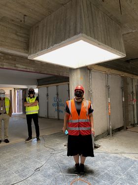 Two people look towards the third person standing in the middle of the picture, facing the camera. She wears a black dress and is under a skylight inside a building under construction