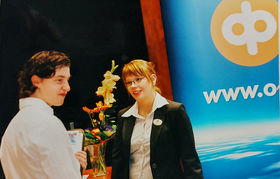 Old photo of two people. They are standing facing each other wearing suits. They are in front of a table with flowers and a poster with OP logo and photo of the sky.