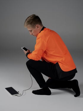 Photo of a person kneeling, facing left, wearing an orange jacket and looking at a mobile phone connected to a solar charger.