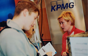 An old photo of two people close up. One is reading a broschure and another has her mouth open. KPMG logo in the background.