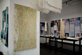 Aalto textiles presented in an exhibition in New York, a sample on the wall, others hanging from the ceiling, and a pictured wall behind in the gallery