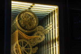 A kinetic light sculpture at night time. Cogs and wheels are golden brown, with an illusion of a continuum into the wall.