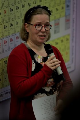 A presenter speaks with a microphone standing in front of a large poster of the periodic table of elements.