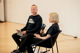 A woman sitting in a chair and talking to a microphone, a man next to her listening