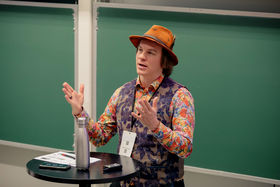 A man in a colorful shirt and a hat standing and speaking 