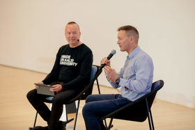 A man sitting in a chair and talking to a microphone, another man next to him listening
