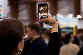 A person is holding a phone and taking a photo with it. You can see a crowd of people in a big hall through the phone screen.