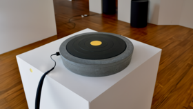 A cylindrical concrete object with a cable plugged into it placed on a white exhibition pedestal