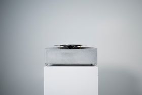 A square concrete block with a metal plate like looking thing on top places on a white exhibition pedestal