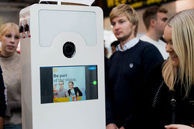 Smiling woman is standing next to a white box that has a screen and a picture of two smiling persons in the photo