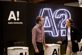 Two people are talking and standing in front of a black wall with logo of Aalto University and a big A? in it