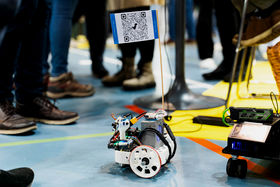 A robot with big wheels and a flag with qr code is on a floor. Feet on the background.