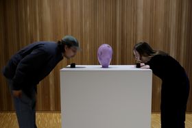 Two people blowing into two black objects on each side of the pink glass object