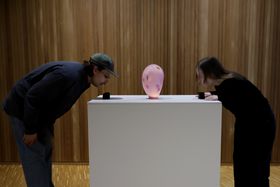 Two people blowing into two black objects on each side of the pink glass object - the objects light up