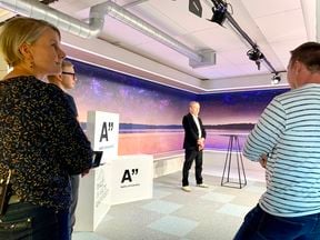 nainen ja kaksi miestä seisovat studiossa kuvan etualalla, taaempana mies odottaa kameran käynnistymistä virtuaalikonferenssissa