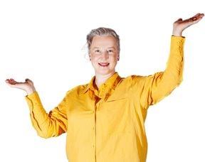 Henkilökuvassa Riikka Mäkikoskela katsoo kameraan, kädet ovat kohotettuina kämmenet ylöspäin, hänellä on yllään keltainen pellavapaita.