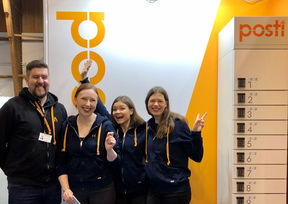 Four people are wearing dark hoodies, standing infront of orange wall with Posti logo, smiling and waving. One is showing a victory sign with fingers.