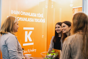 Four people standing infront of orange-colored walls with texts and K group logo. A bowl of candy.