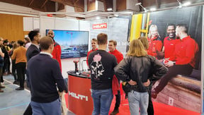 People are standing in front of a wall with huge picture of people with red shirts sitting on a coffee break and a Hilti logo. People are talking with eachoterh.